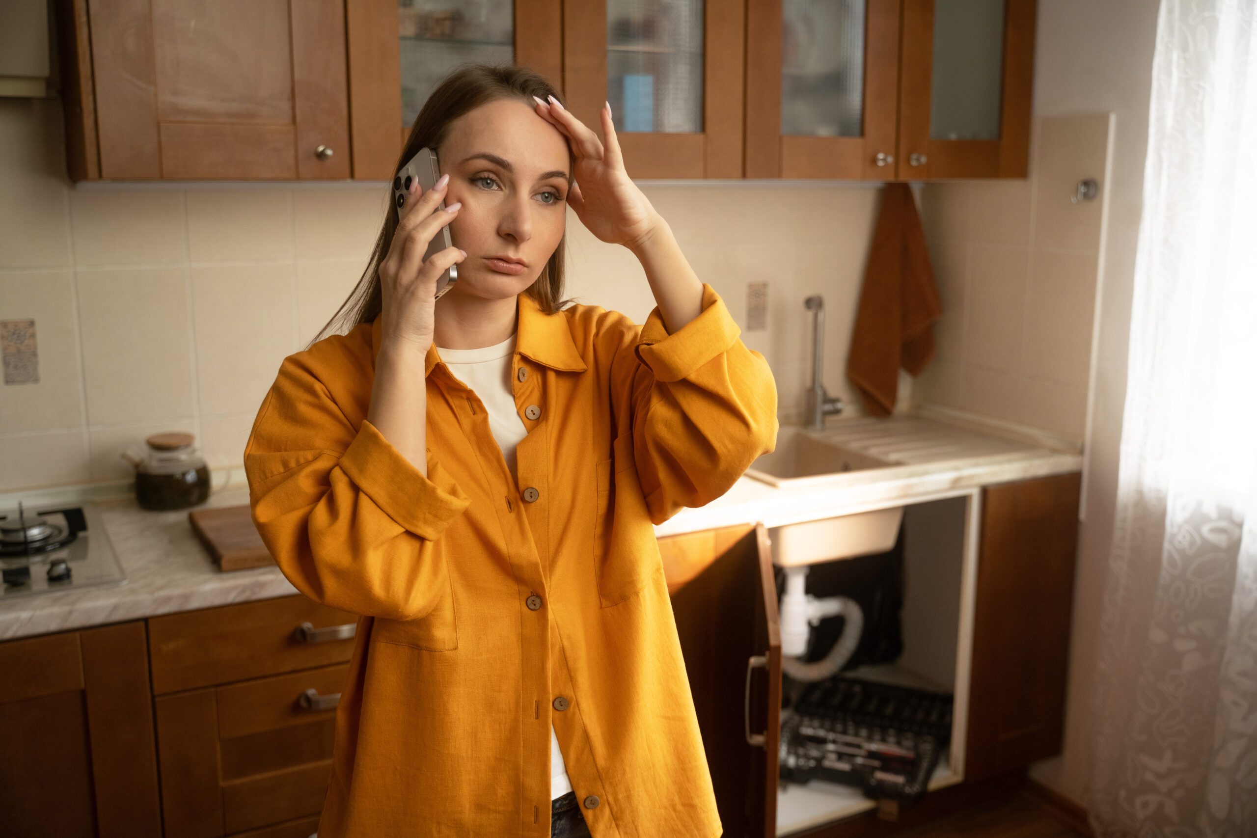 A woman in a yellow shirt stands in her kitchen while discussing urgent plumbing issues with a repair service regarding a leaky sink that needs immediate attention.